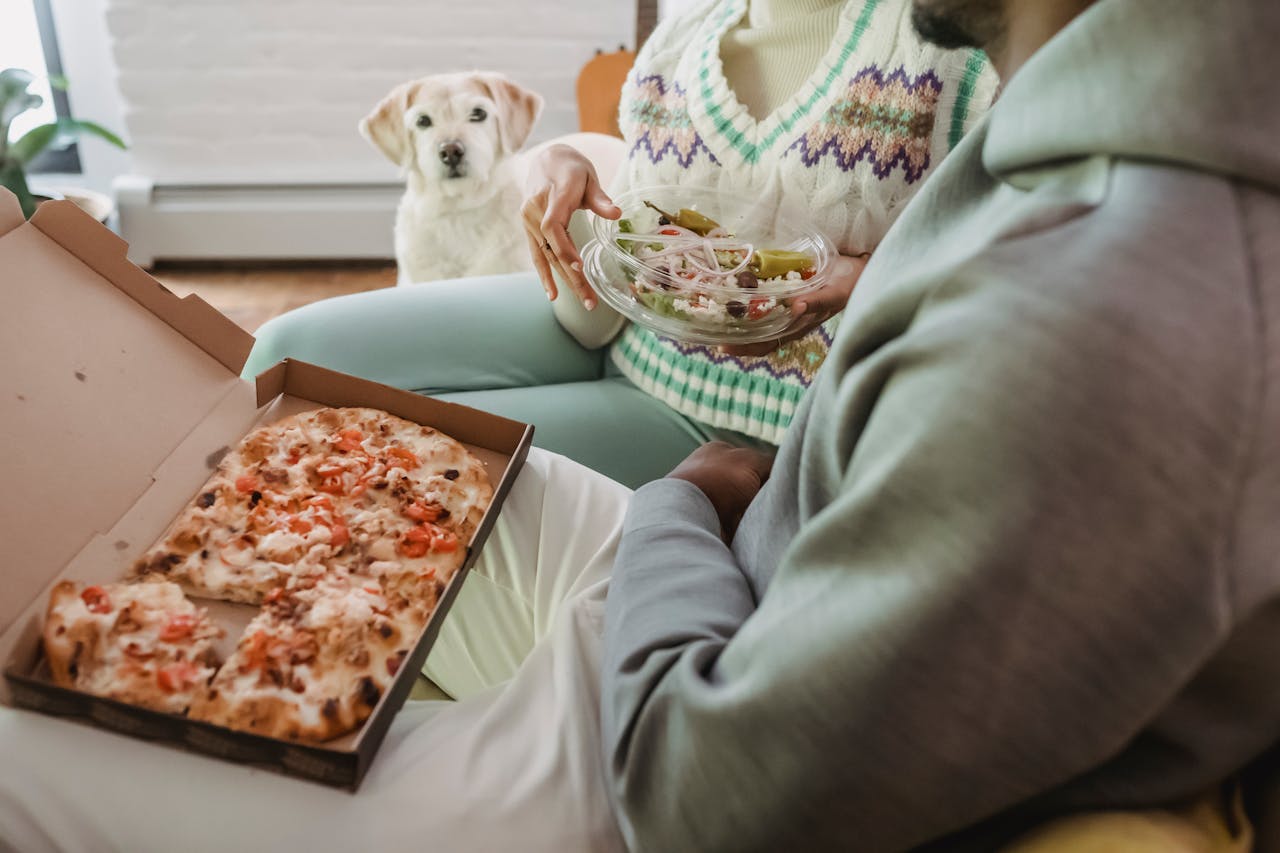 Egy nő és egy férfi ül a kanapén, pizzát eszik, ami káros étel a kutyáknak és a kutyájuk figyeli őket.