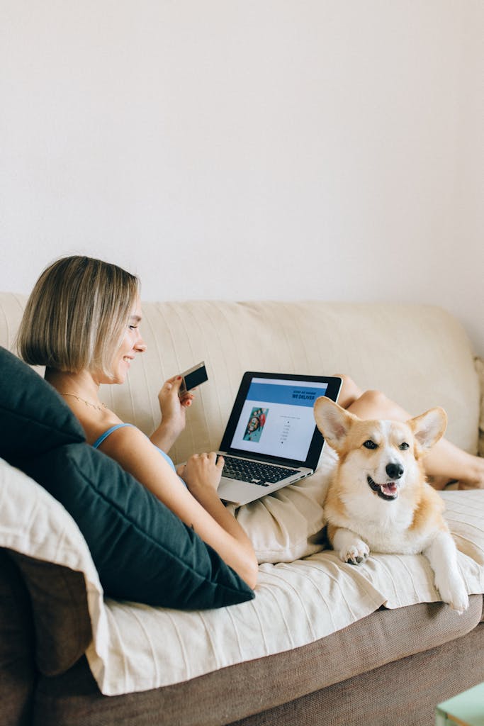 Woman with laptop and corgi dog shopping online with credit card on sofa.